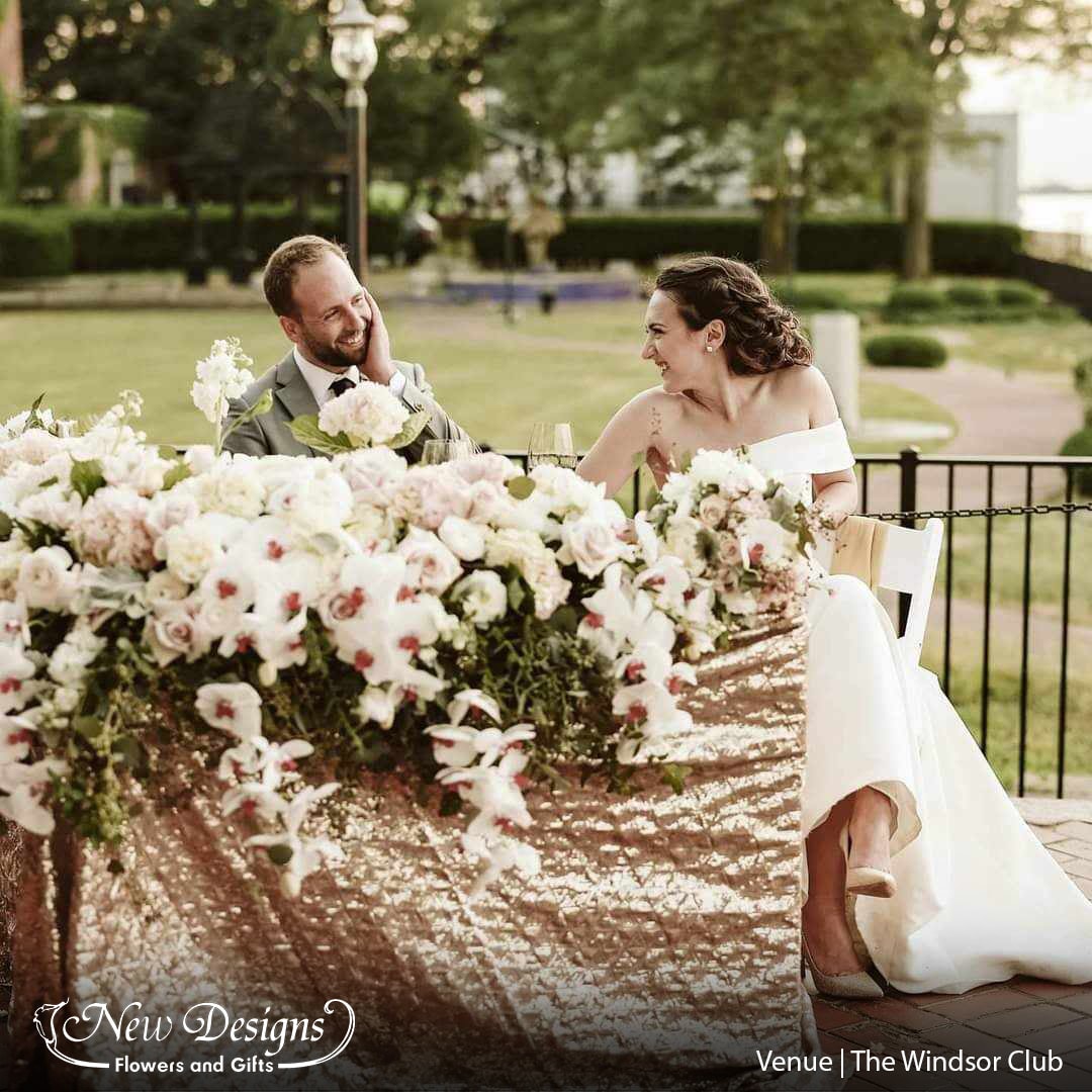 A bride and groom sit at a head table lined with a gold sequined tablecloth overflowing with white roses and orchids at a waterfront venue